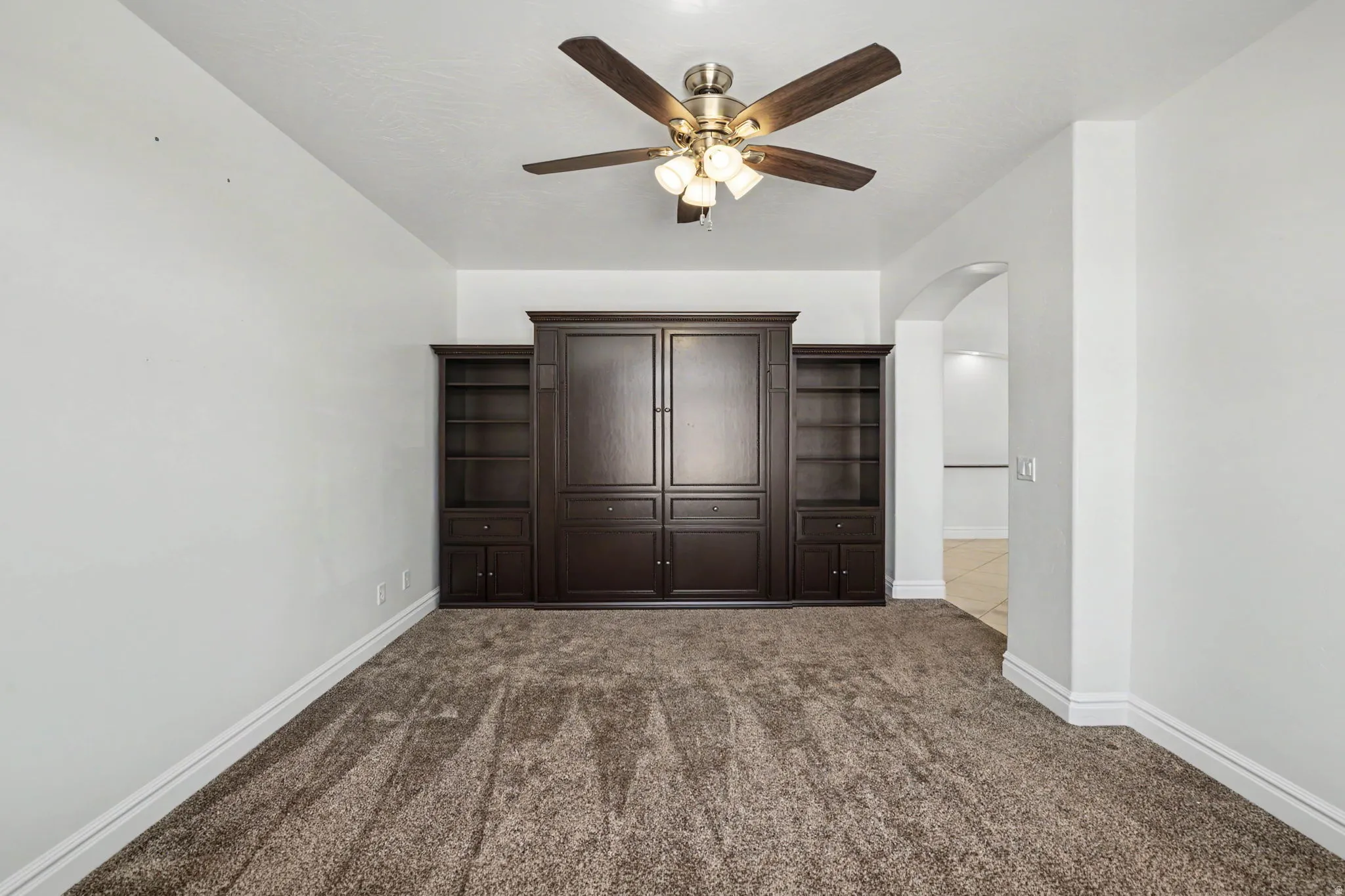 Built-in wall unit with shelving and cabinetry, ceiling fan with integrated lighting, arched doorway, and carpeted flooring