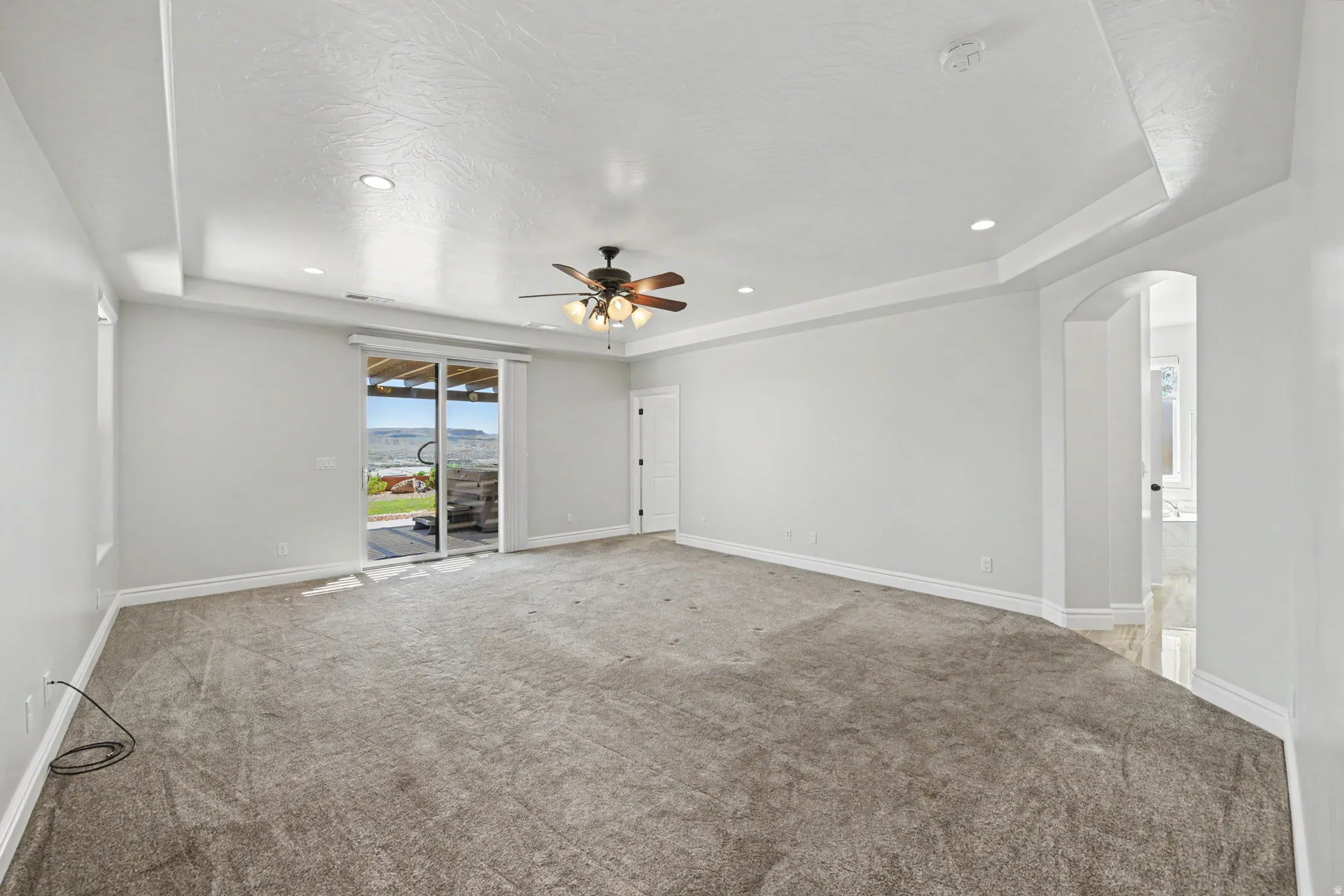 Expansive carpeted room featuring a tray ceiling with recessed lighting and a ceiling fan, sliding glass door access to an outdoor area, and an arched doorway
