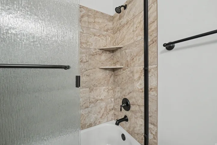 Bathtub shower with textured glass enclosure, tiled surround, built-in corner shelves, and matte black fixtures