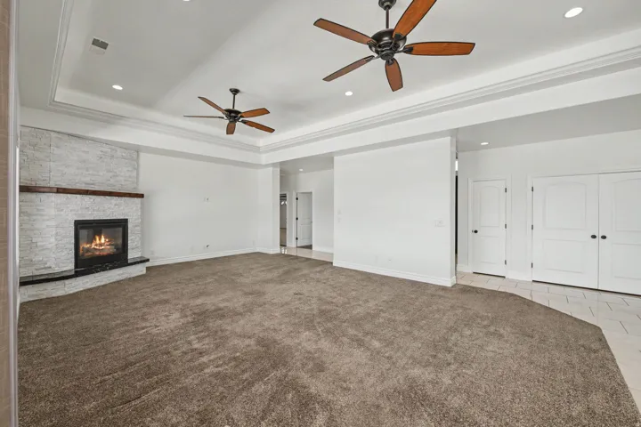 Spacious room featuring a stacked stone fireplace with a dark wood mantel and hearth, tray ceiling with recessed lighting, two ceiling fans, and neutral carpeting