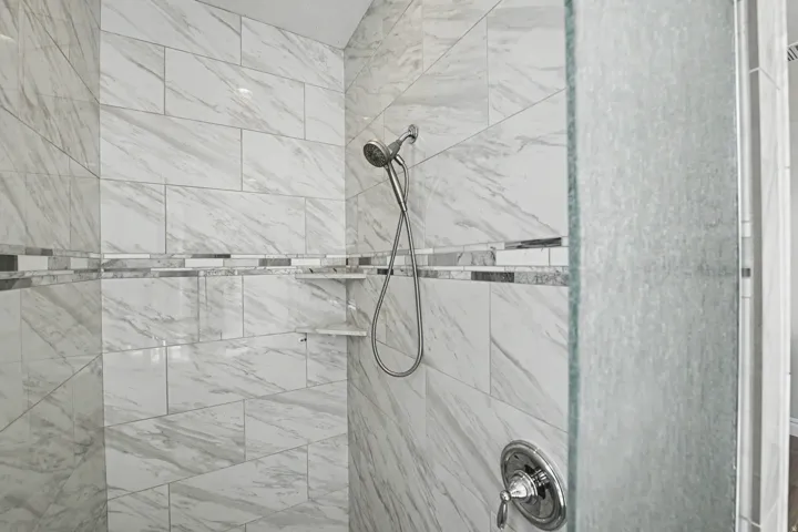 Shower enclosure featuring large format white tile with gray veining, decorative mosaic tile band, built-in corner shelving, and a chrome shower fixture