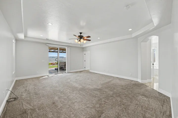 Expansive carpeted room featuring a tray ceiling with recessed lighting and a ceiling fan, sliding glass door access to an outdoor area, and an arched doorway