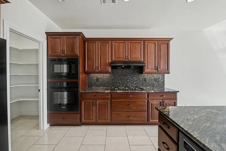 Built-in wall oven and microwave, dark wood cabinetry with crown molding, gas cooktop with coordinating backsplash, stone-finish countertops, and light-toned tile flooring