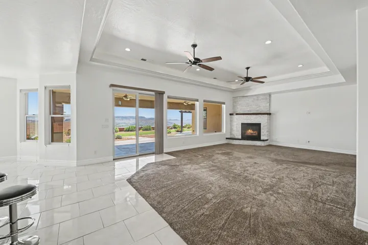 Tray ceiling with recessed lighting, dual ceiling fans, stone-clad fireplace with wood mantle, large sliding glass doors, and glossy tile flooring