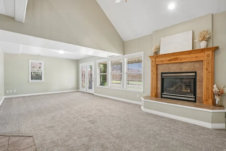 Vaulted ceiling, carpeted flooring, a fireplace with a wood mantel and tile surround, multiple windows, and glass patio doors