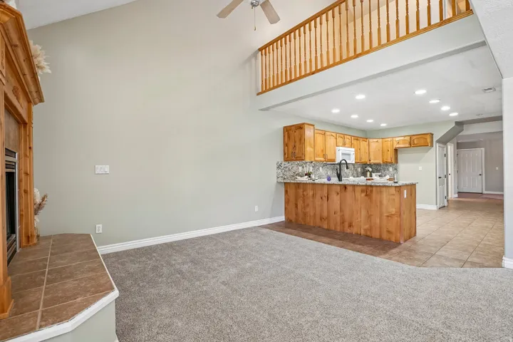 Vaulted ceiling with ceiling fan, wood-finish mantel and tile hearth, kitchen with wood cabinetry and stone countertops, tile flooring, and carpeted living area