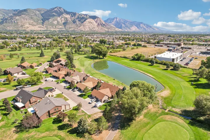 Aerial perspective showcasing a residential area with varied rooflines, a prominent pond, expansive green spaces, and a mountainous background