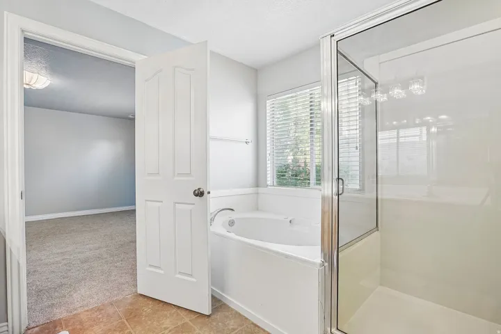 White soaking tub, glass-enclosed shower, window with blinds, and tile flooring