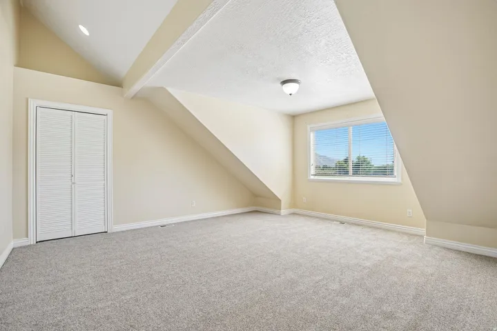 Spacious room featuring sloped ceilings, a large window with blinds, soft gray carpet, white louvered bifold closet doors, and recessed lighting