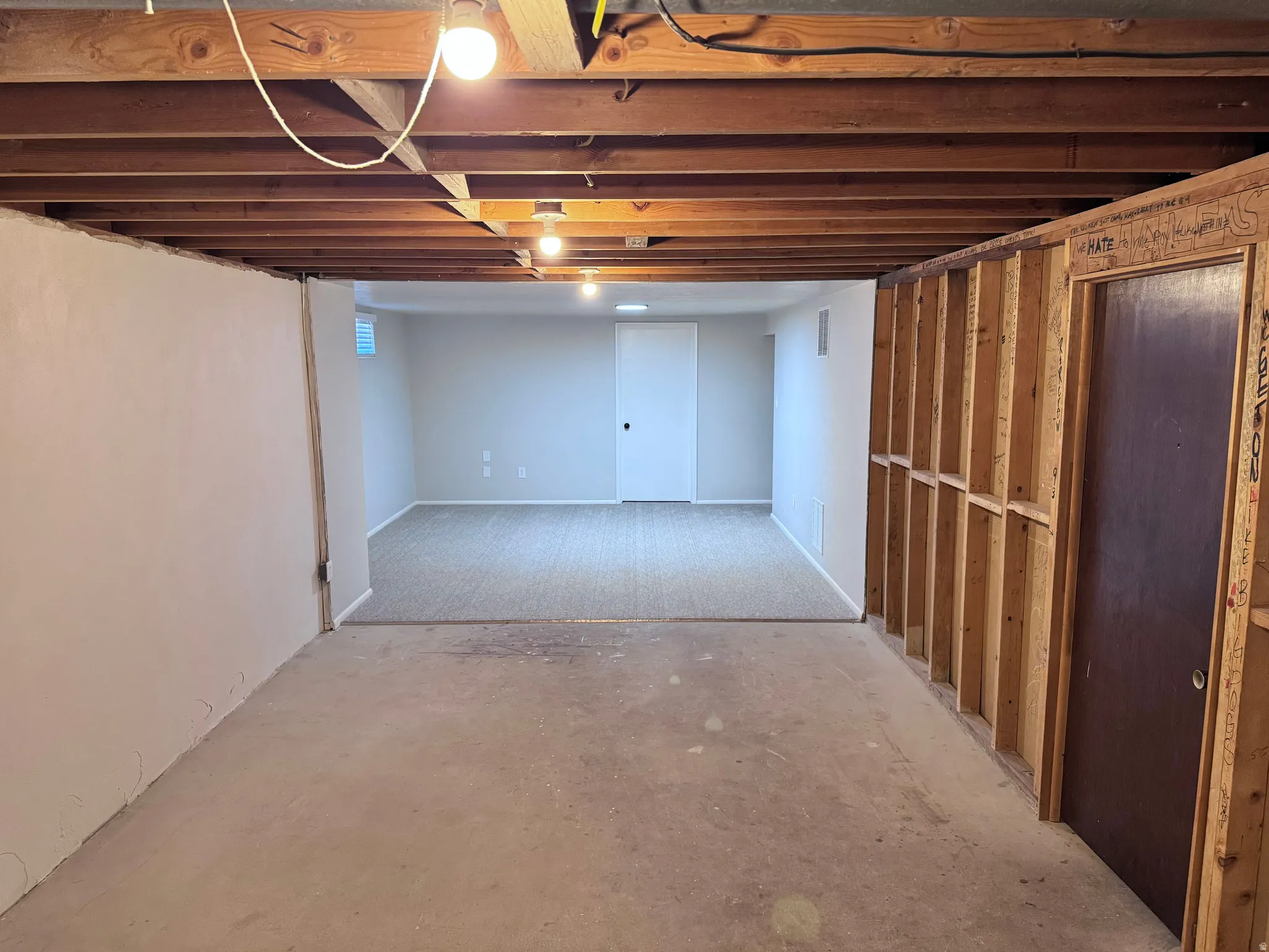 Expansive basement space featuring exposed wood joists, partial drywall, framed wall sections, and a finished area with carpeting and light gray walls