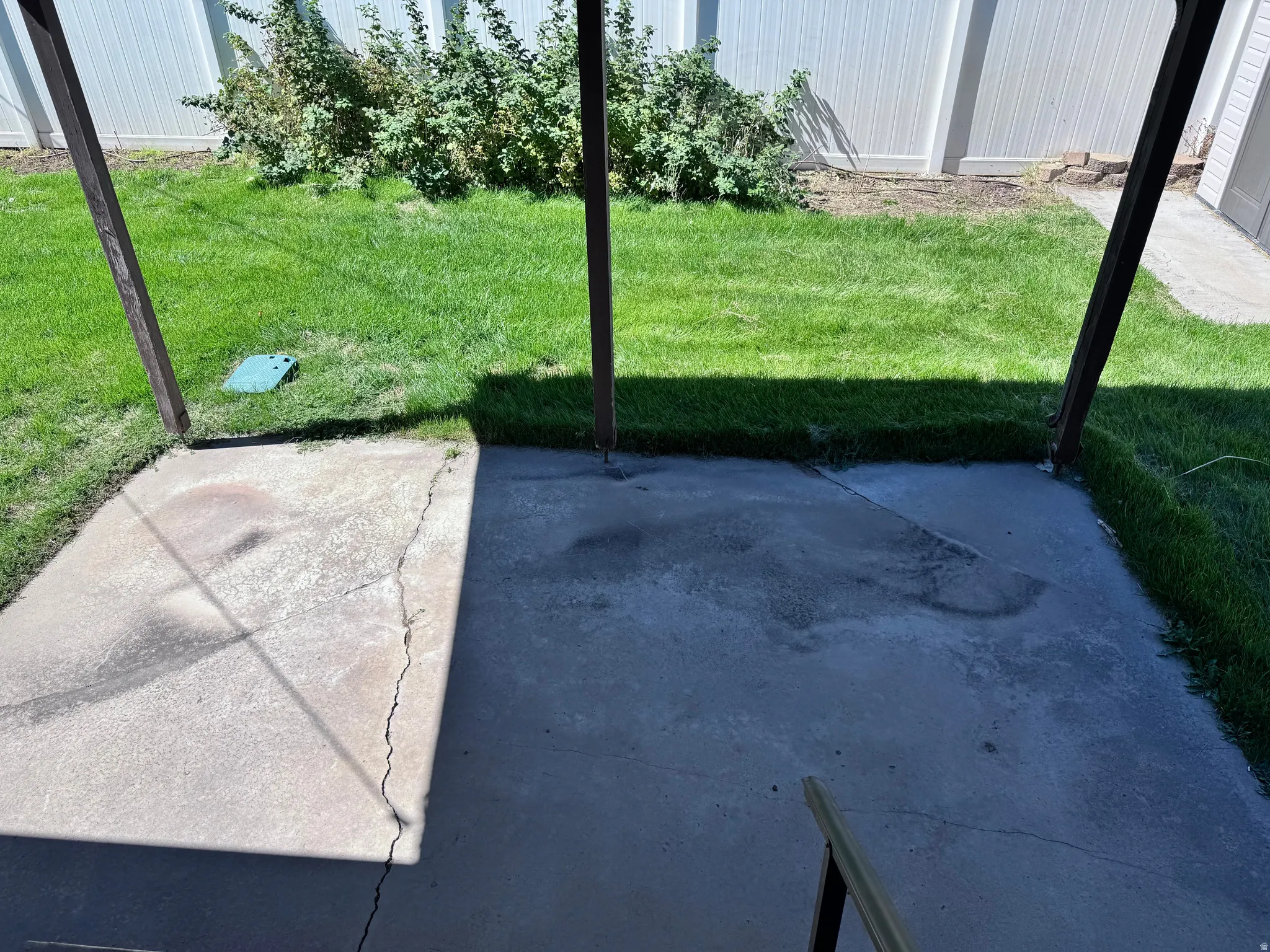 Concrete patio with overhead structural supports, bordered by a green lawn and mature shrubbery, enclosed by a privacy fence