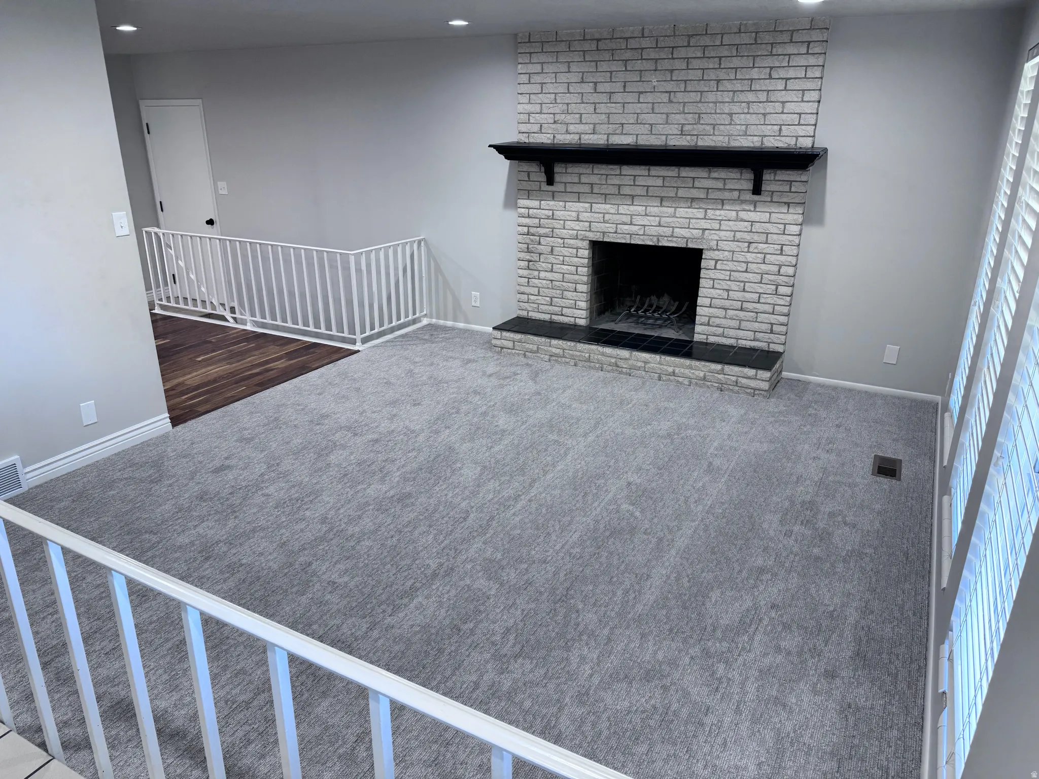 Living space featuring a brick fireplace with a black mantel, gray carpeting, a white railing, recessed lighting, and wood-finish flooring