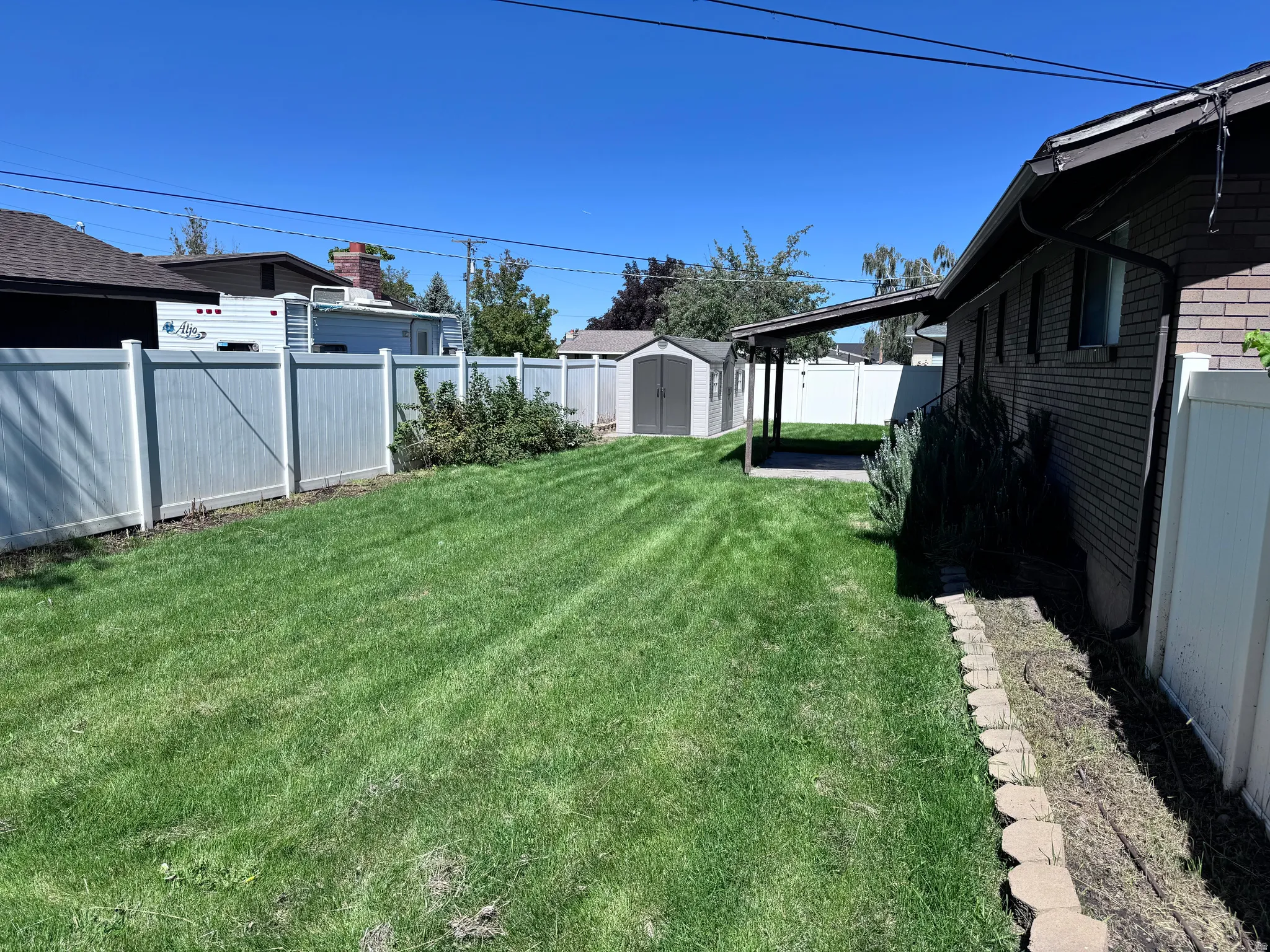 Expansive green lawn with a white privacy fence, brick exterior, covered patio, and storage shed