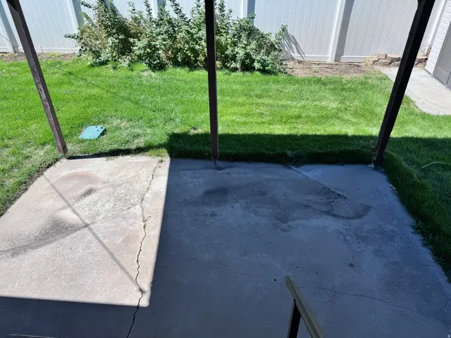 Concrete patio with overhead structural supports, bordered by a green lawn and mature shrubbery, enclosed by a privacy fence