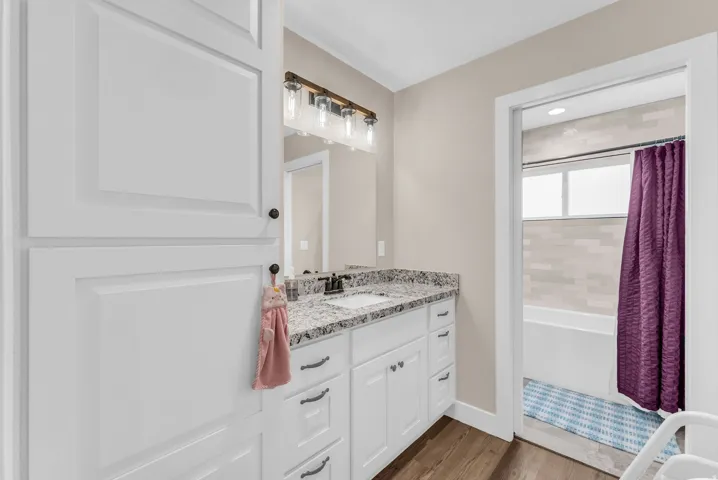 White shaker-style cabinetry with dark hardware, granite-look countertop, dark wood-finish flooring, and a tiled shower surround
