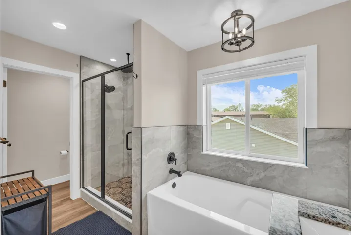Glass-enclosed shower with dark fixtures and tile surround, separate soaking tub with tile wainscoting, and wood-finish flooring