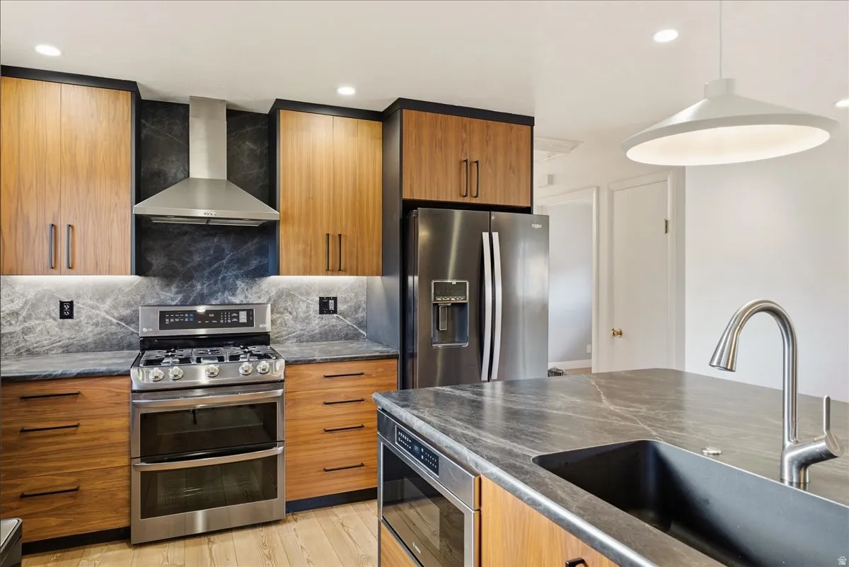 Modern kitchen with wood-finish cabinetry, stainless steel appliances, dark stone countertops, and a large sink with a gooseneck faucet