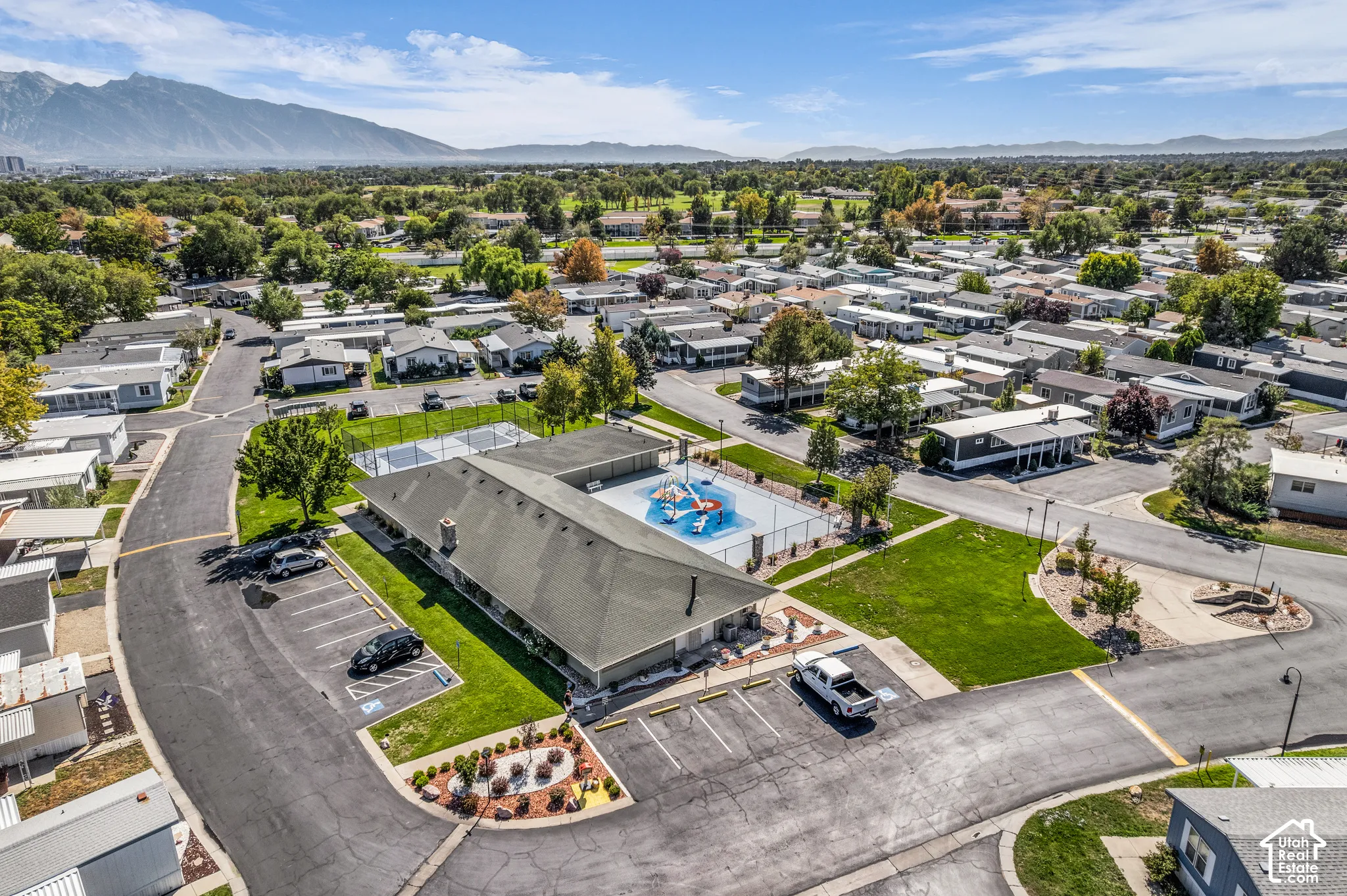Community grounds featuring a large building with a dark roof, a fenced swimming pool, a separate fenced court, and paved parking areas