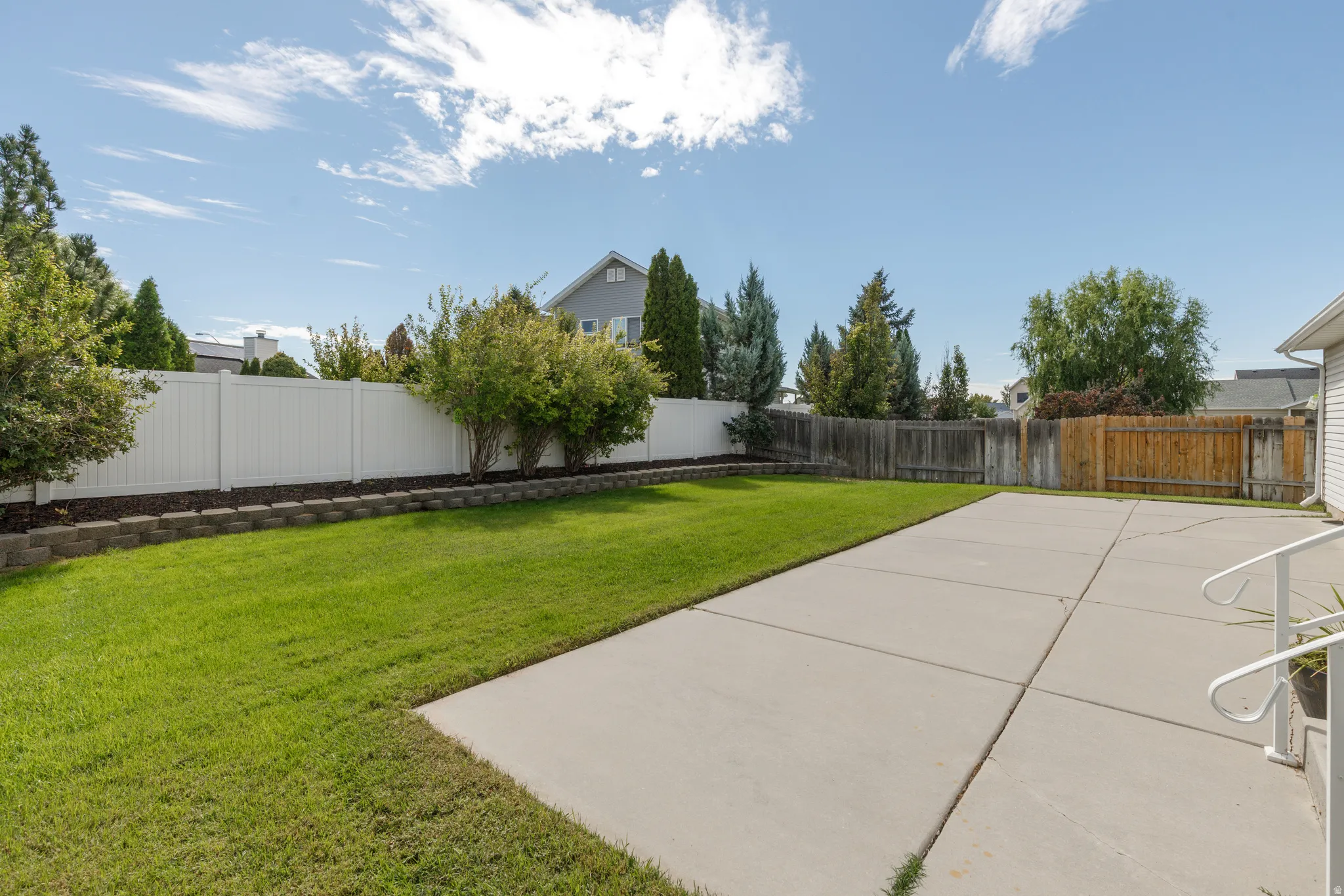 Expansive concrete patio, manicured lawn, established shrubs, and white privacy fencing