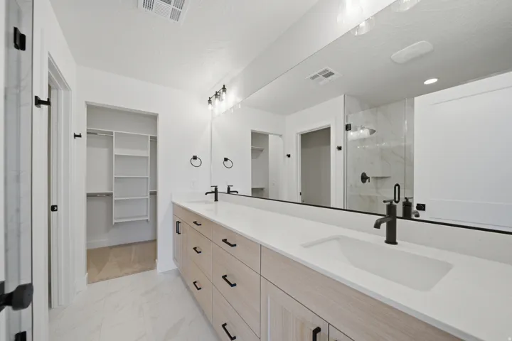 Dual vanity with integrated sinks, light wood-finish cabinetry, a full-width mirror, a glass-enclosed shower, and a walk-in closet with built-in shelving