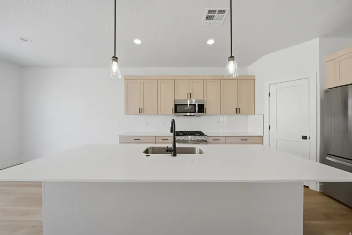 Spacious kitchen island with an undermount sink and matte black faucet, complemented by light wood-finish cabinetry, stainless steel appliances, and a tiled backsplash