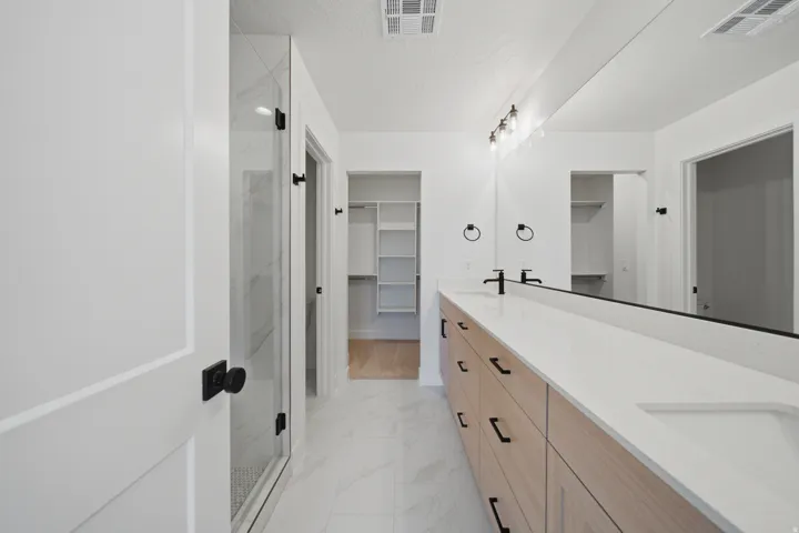 Dual vanity with integrated sinks, light wood-finish cabinetry, matte black fixtures, an expansive mirror, and a glass-enclosed shower with tile surround