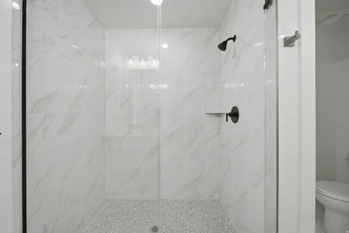 Walk-in shower featuring glass enclosure, marble-look tile walls, a built-in shelf, and matte black fixtures