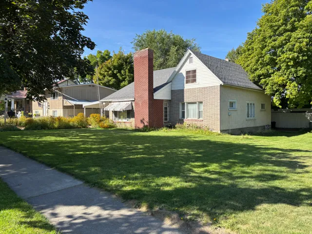 Exterior featuring brick and siding finishes, a prominent red brick chimney, an awning over a window, a partial privacy fence, and mature trees providing shade