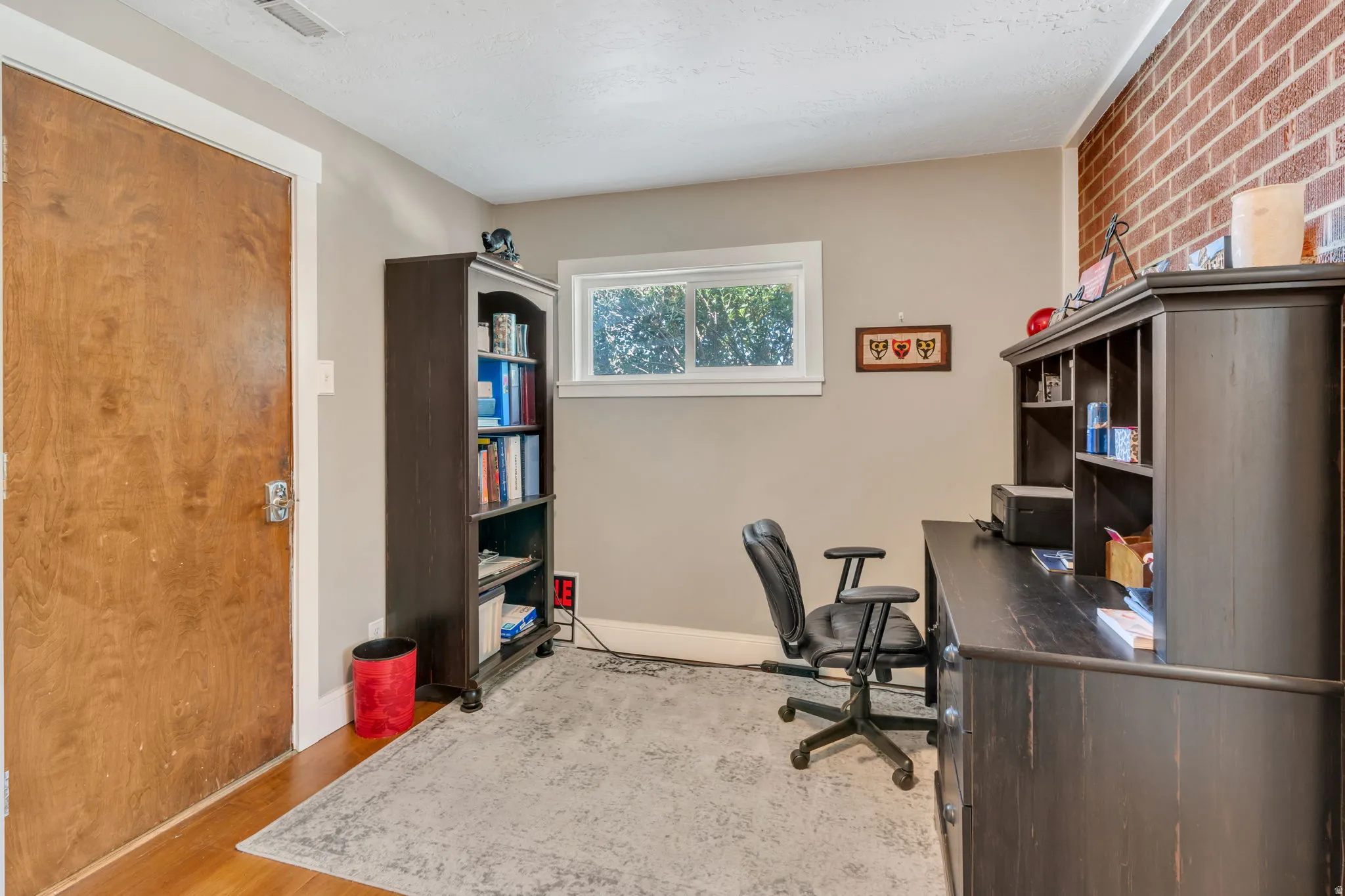 Wood-finish flooring, a wood-panel door, a window with white trim, and an exposed brick accent wall
