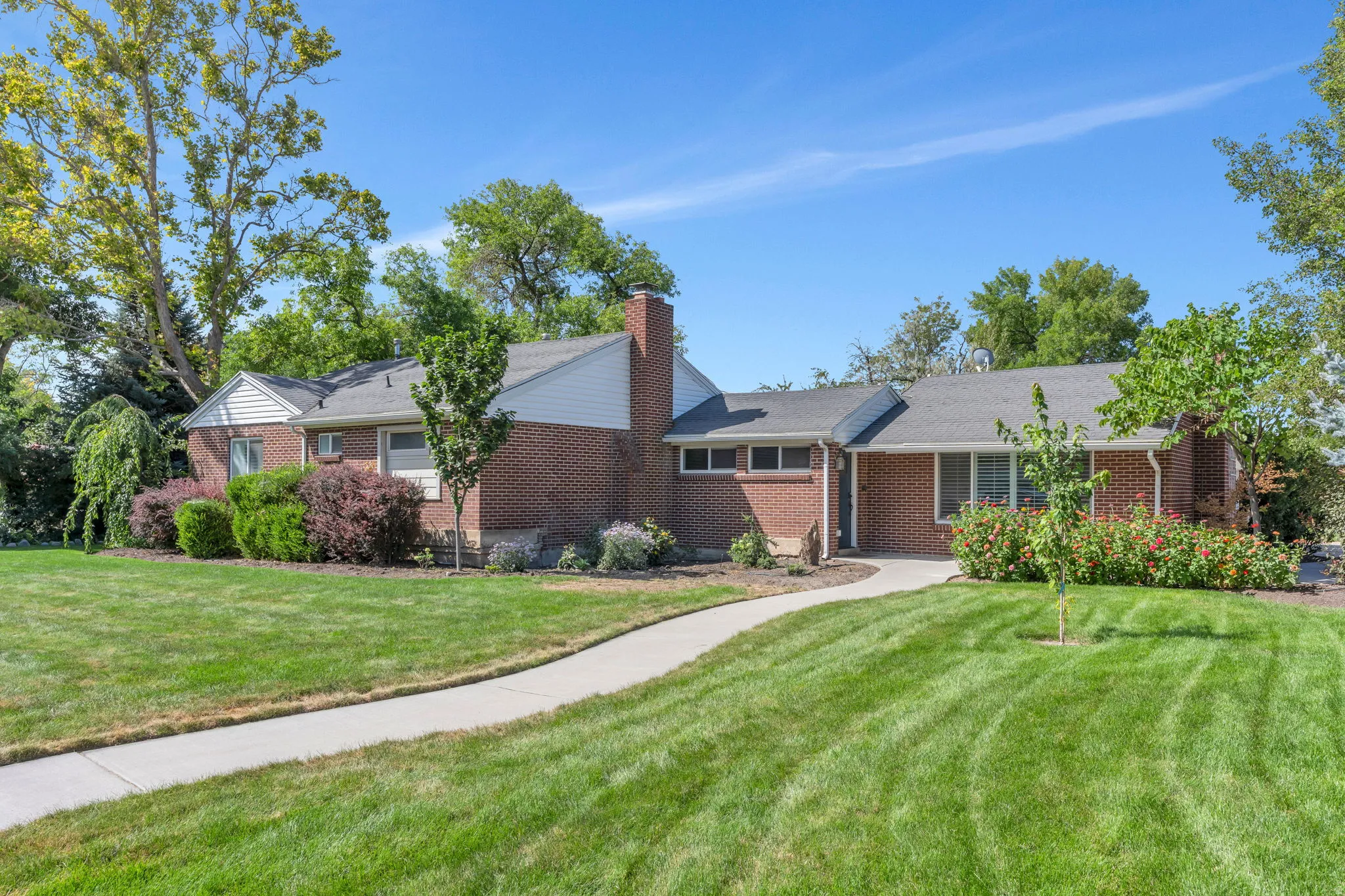 Brick exterior with white siding accents, mature trees, established landscaping, a concrete walkway, and a spacious lawn
