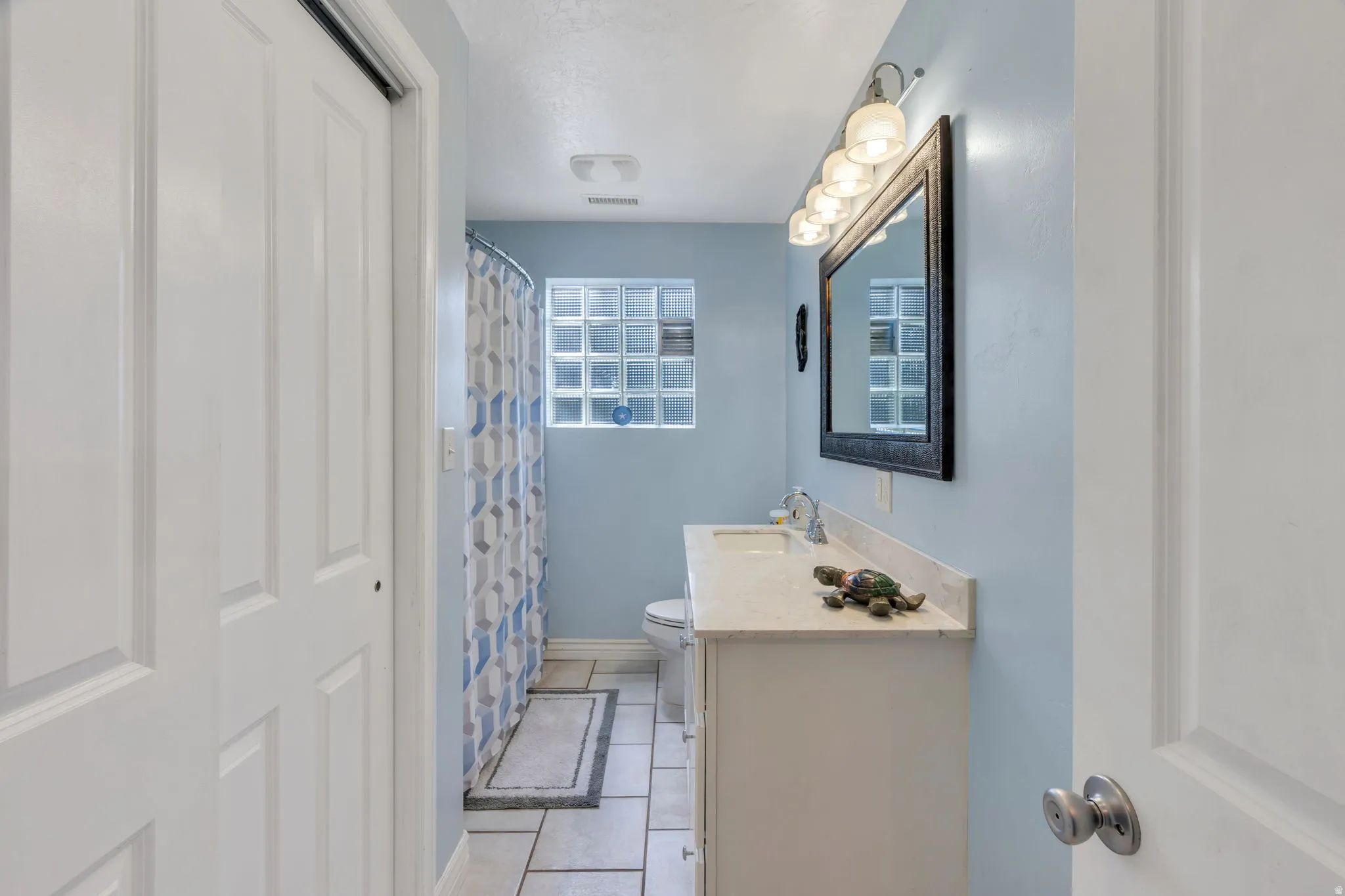 White paneled bifold closet doors, light blue painted walls, rectangular tile flooring, glass block window, and a white vanity with a light-colored countertop