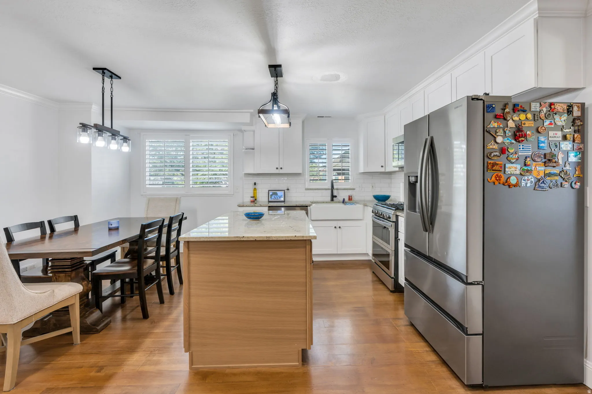Hardwood flooring throughout, white shaker cabinetry with crown molding, stainless steel appliances, central island with stone countertop, and plantation shutters