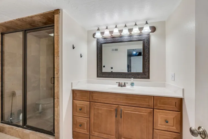Glass-enclosed shower with tile surround, wood-finish vanity with countertop, framed mirror, and a 5-light fixture