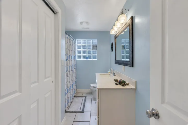 White paneled bifold closet doors, light blue painted walls, rectangular tile flooring, glass block window, and a white vanity with a light-colored countertop
