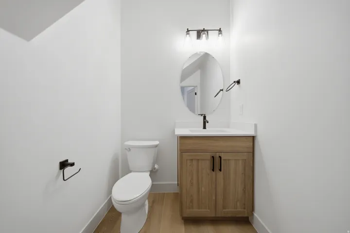 Wood-finish flooring, light wood vanity, white countertop, oval mirror, and dark-finish fixtures