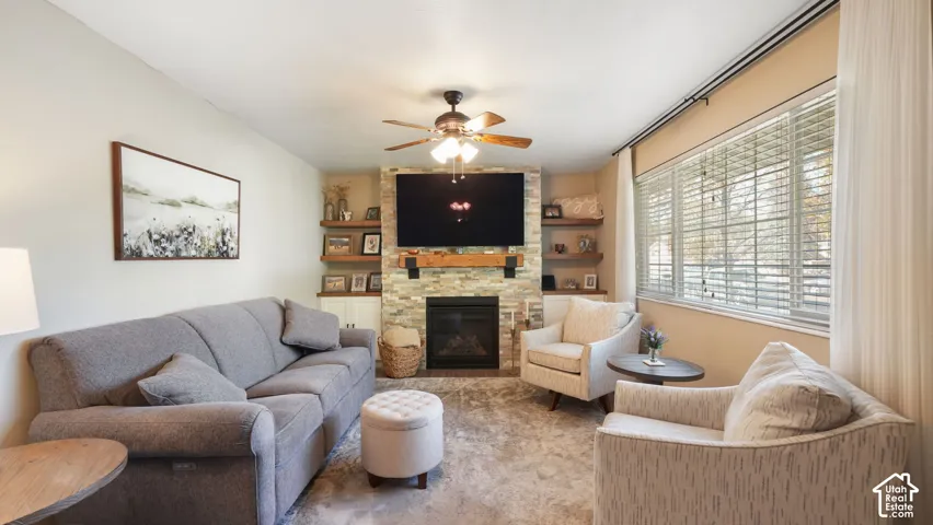 Living area featuring a stone-clad fireplace with wood mantel, built-in cabinetry, wood-finish open shelving, and a ceiling fan with integrated lighting
