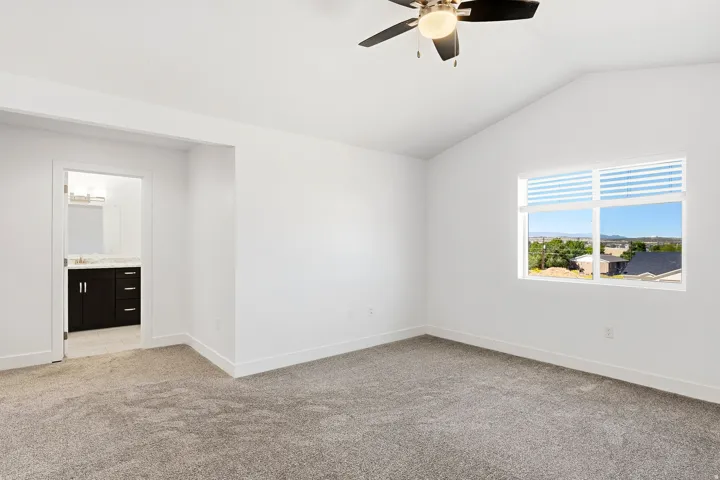 Vaulted ceiling with an attached ceiling fan, plush carpet flooring, a large window with horizontal blinds, and an integrated bathroom featuring a dark wood-finish vanity with a light countertop