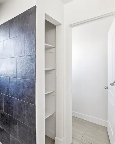Built-in shelving unit, dark large-format wall tiles, white trim, light-toned tile flooring, and a white paneled door with a brushed metal handle