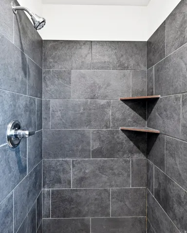 Shower with large format gray wall tiling, chrome shower head, chrome control knob, and two corner storage shelves