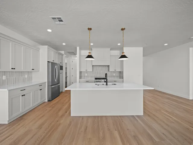 Kitchen featuring wood-finish flooring, an oversized island with a built-in sink, stainless steel refrigerator, white cabinetry, and pendant lighting