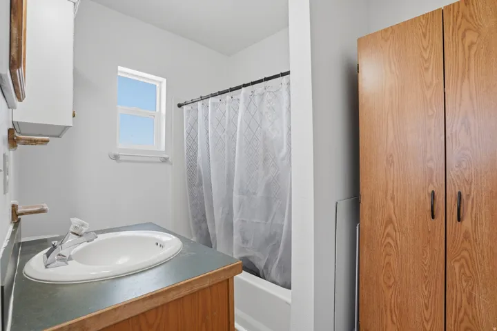 Built-in wood-finish cabinetry, single-basin sink with chrome faucet, white bathtub with shower curtain, and a window with natural light