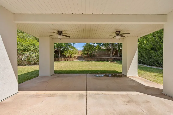 Covered patio with a beadboard ceiling, dual ceiling fans, concrete flooring, and a terraced backyard featuring a manicured lawn and retaining walls