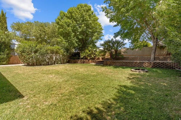 Expansive backyard featuring a green lawn, tiered block retaining walls, mature trees, and a built-in fire pit