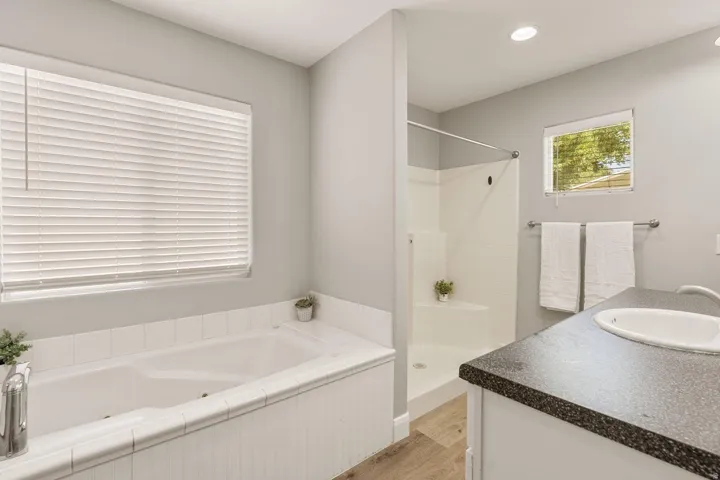 Soaking tub, walk-in shower, single vanity with dark countertop, wood-finish flooring, and recessed lighting