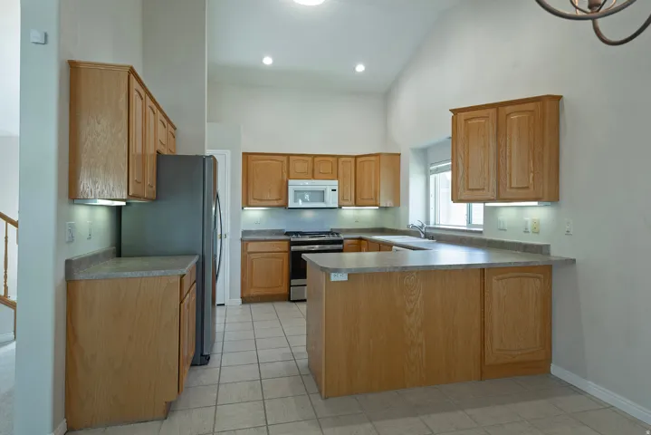 Kitchen featuring vaulted ceilings, wood cabinetry, stainless steel appliances, tiled flooring, and under-cabinet lighting