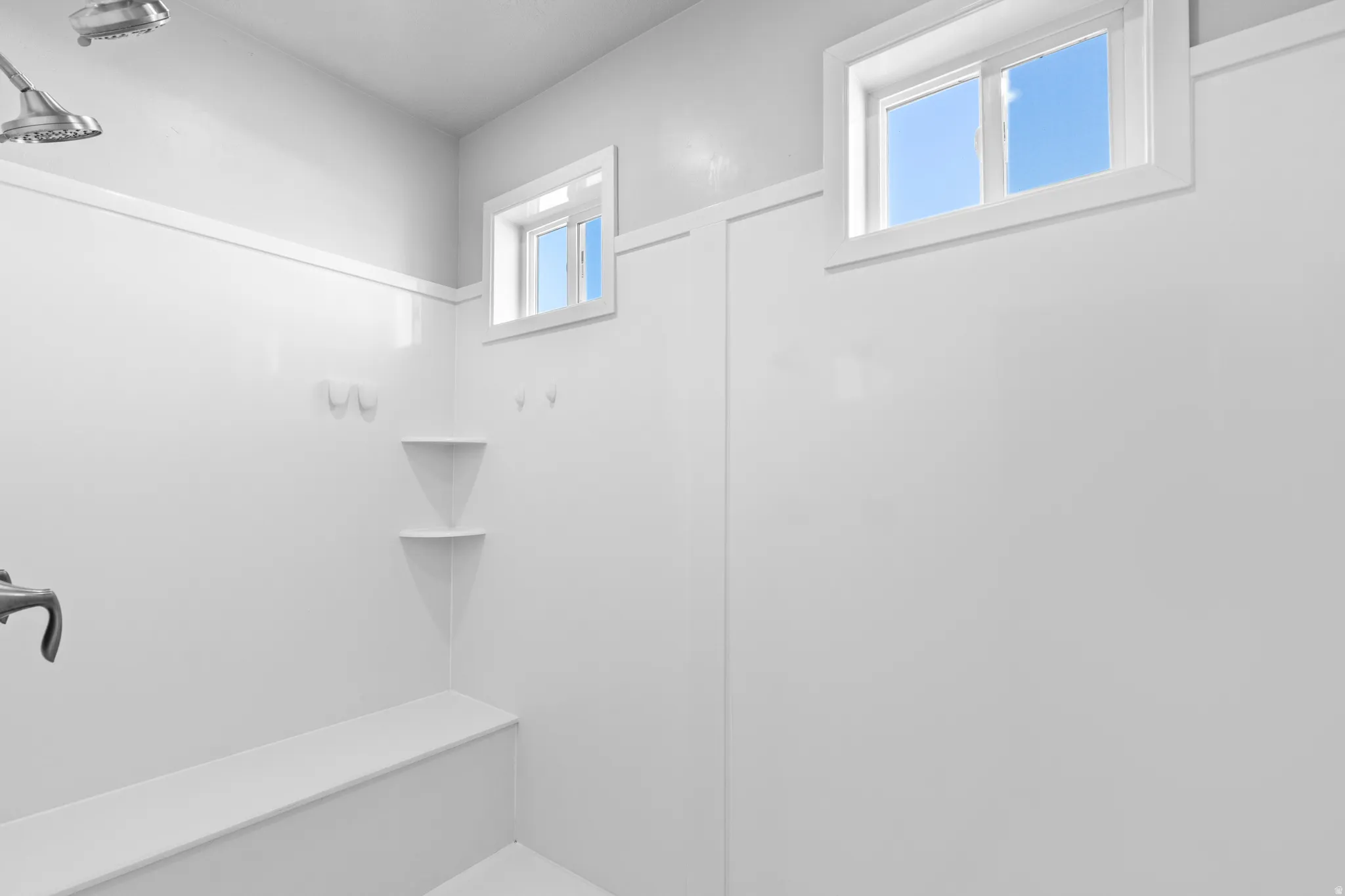 Bright shower enclosure featuring two windows, dual shower heads, a built-in bench, and integrated corner shelving