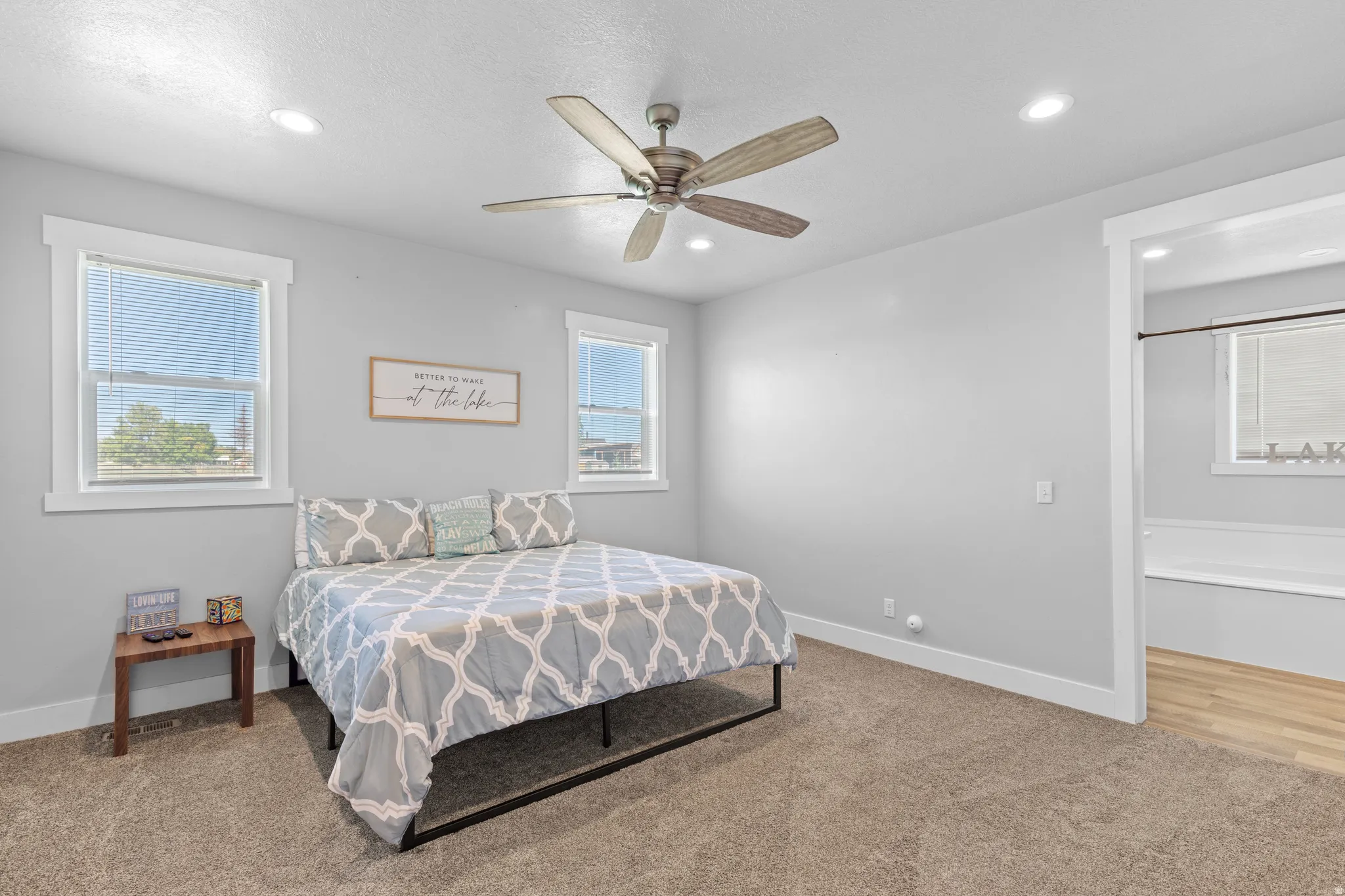 Recessed lighting, ceiling fan, two windows, wall-to-wall carpeting, and an adjacent bathroom with wood-finish flooring and a built-in tub