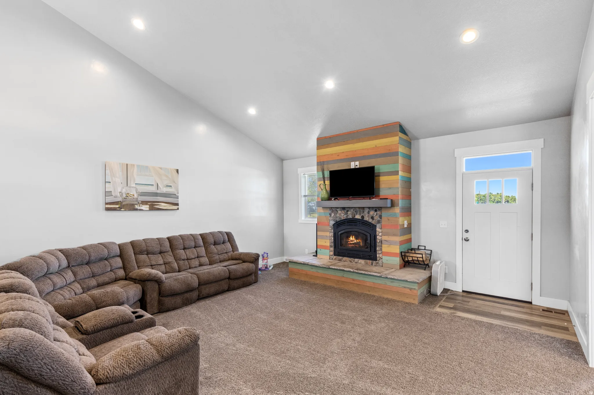 Vaulted ceiling, recessed lighting, wood-finish flooring at entry, white paneled entry door with transom and clear glass inserts, and a colorful wood-clad fireplace with a stone hearth