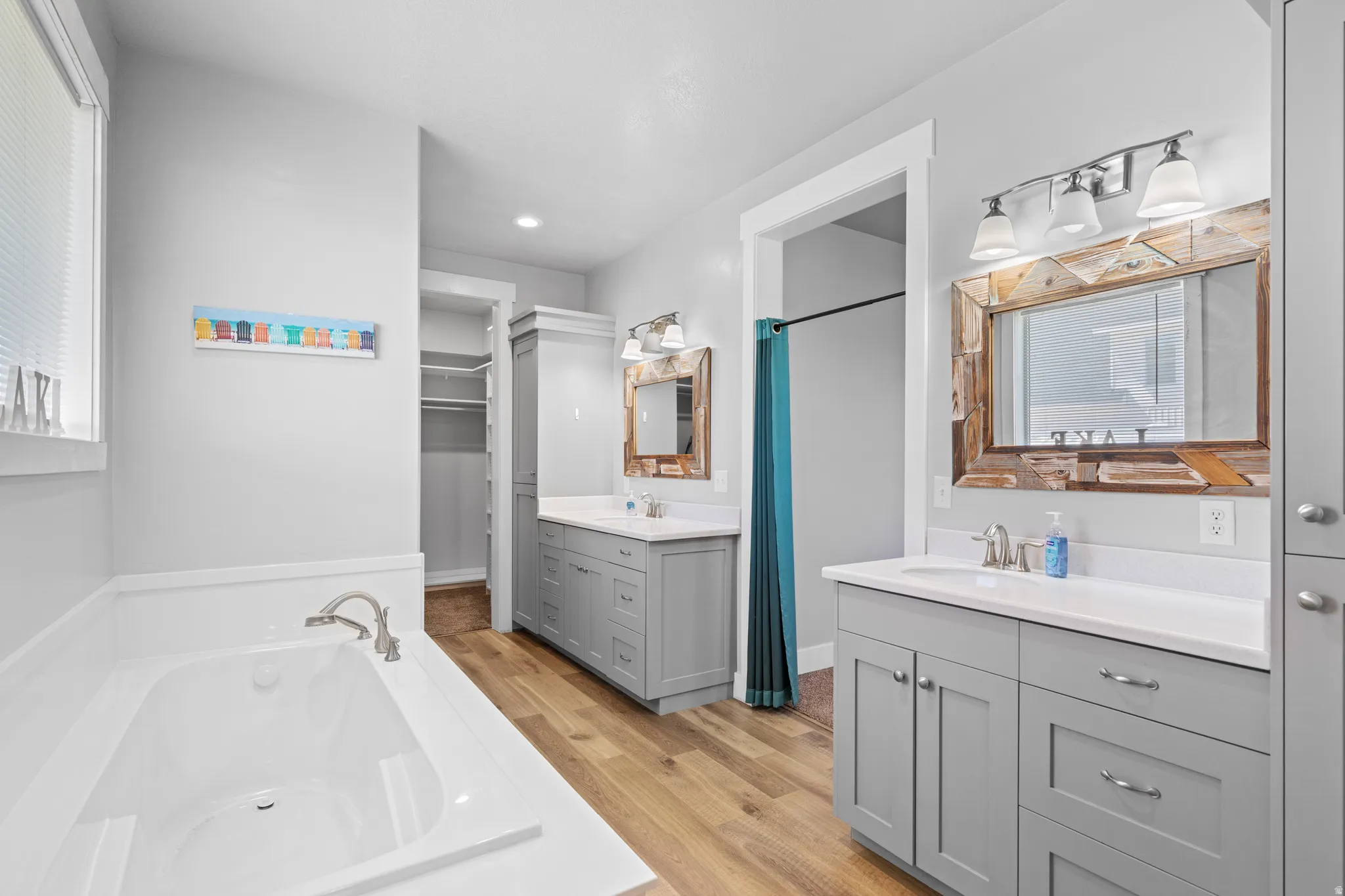 Soaking tub with deck-mounted faucet, dual vanity with shaker-style cabinetry and undermount sinks, wood-finish flooring, and a walk-in closet