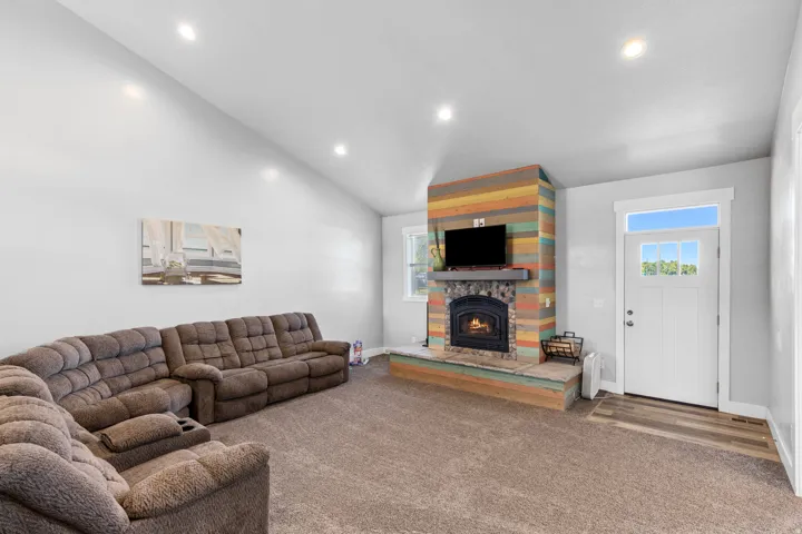 Vaulted ceiling, recessed lighting, wood-finish flooring at entry, white paneled entry door with transom and clear glass inserts, and a colorful wood-clad fireplace with a stone hearth